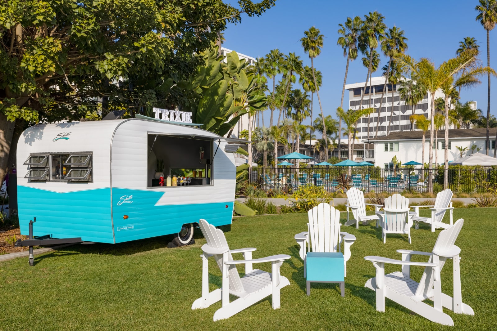 Teal vintage camper trailer on a lawn with white and blue Adirondack chairs in front