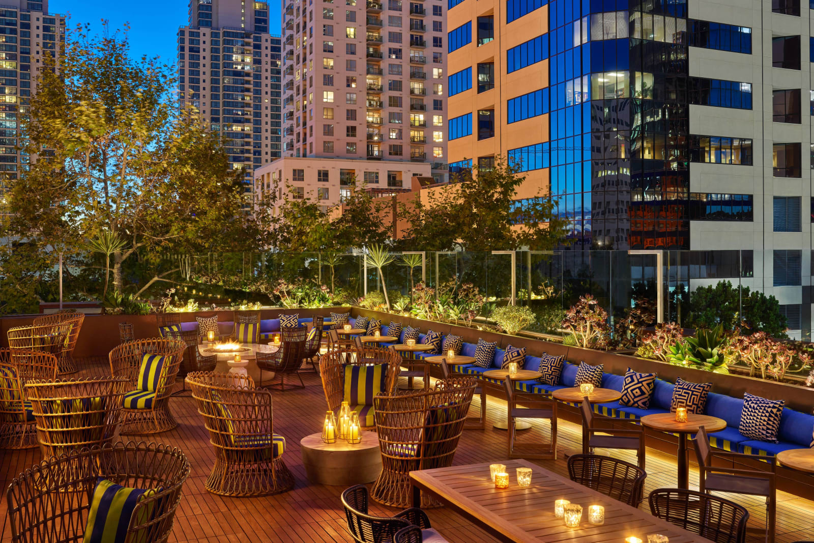 Rooftop terrace at dusk with fire pits, wicker seating and planting, taller office towers behind