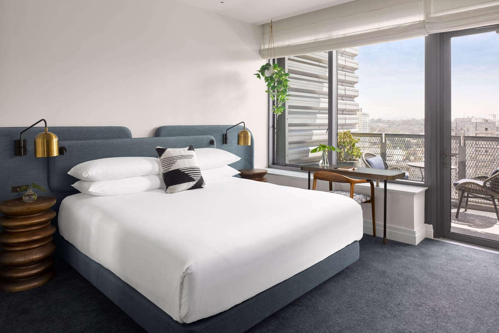 Guest room with a king bed and deep blue headboard, full height glass on the right opening onto a railed balcony above the city