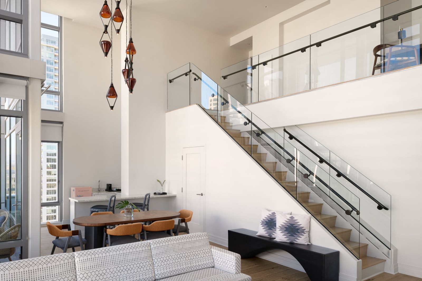 Double height white interior with a staircase behind glass balustrades leading to an upper level and long pendant lights hanging down