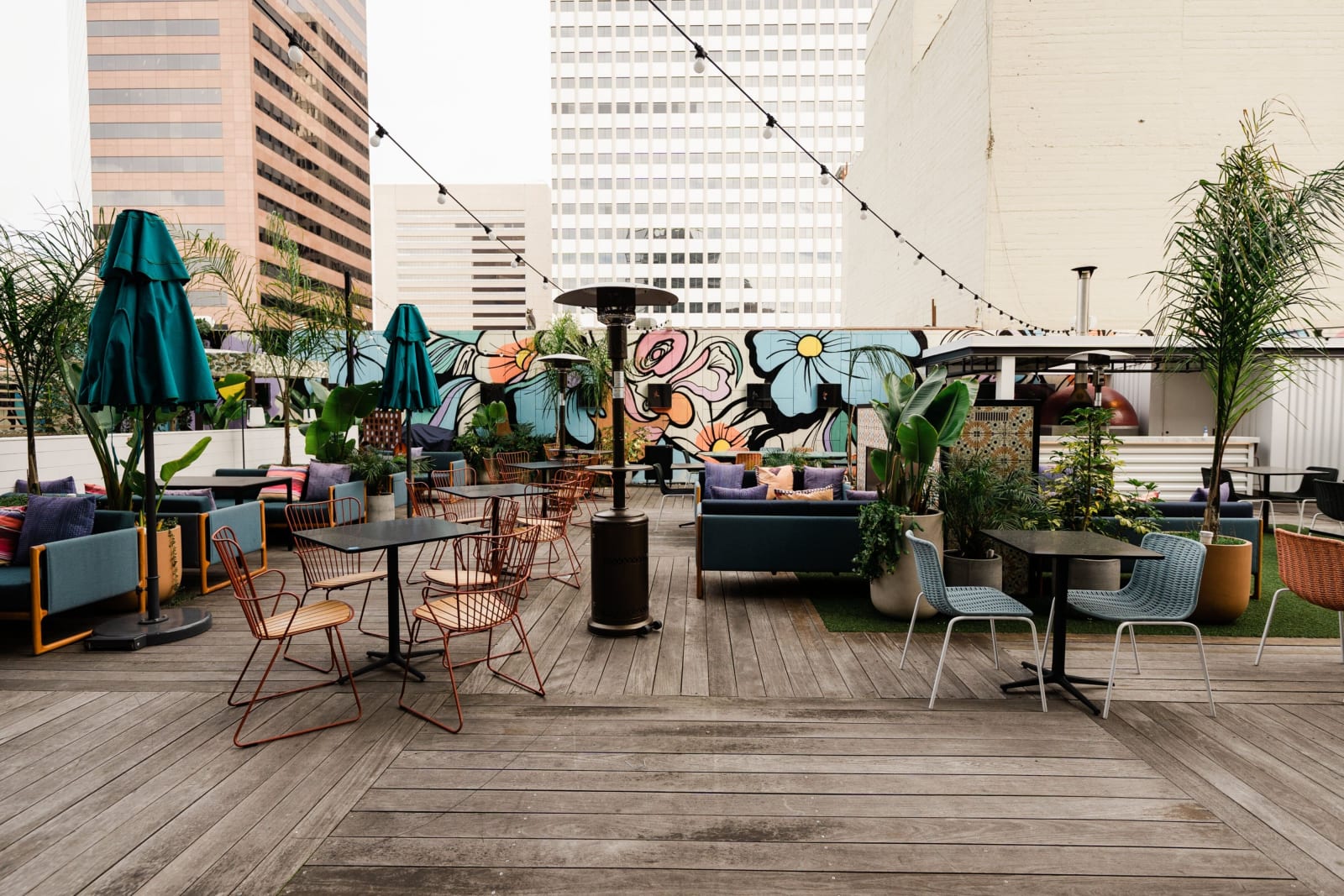 Rooftop terrace at dusk with umbrellas folded, string lights strung between murals and planting, and lit office towers behind