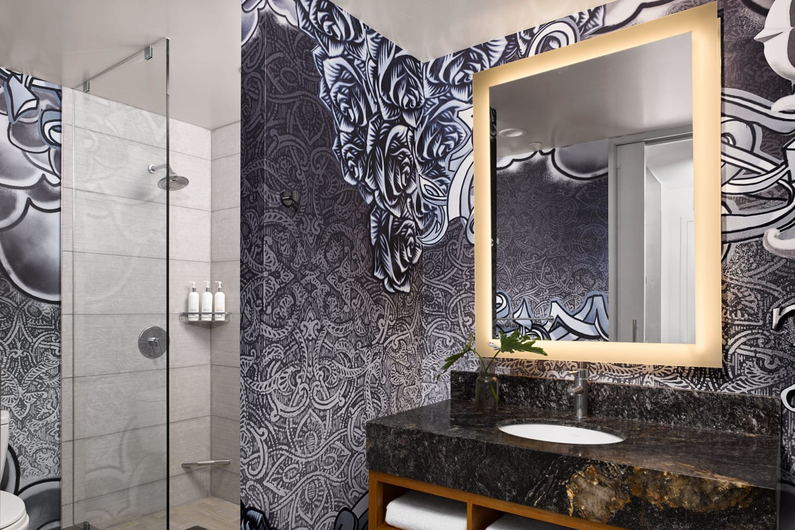 Corner of a bathroom with a walk in shower behind a fixed glass panel on the left and a bold black and white patterned wall with a gold framed mirror on the right