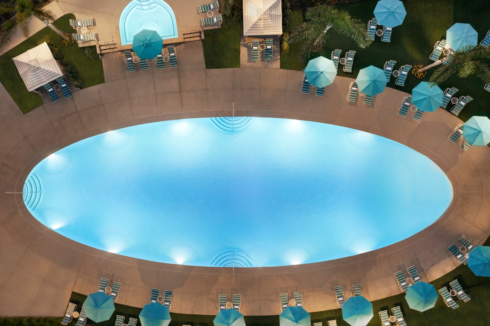 Oval pool lit at night ringed by teal umbrellas and loungers