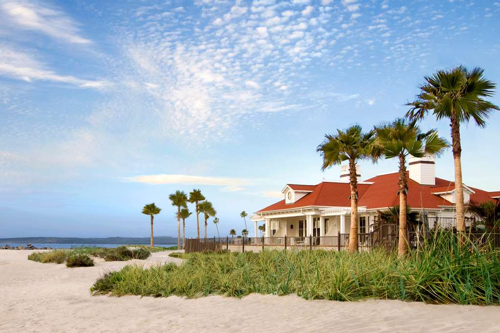 Red tiled white building on the sand with low scrub and palms in front under an open sky
