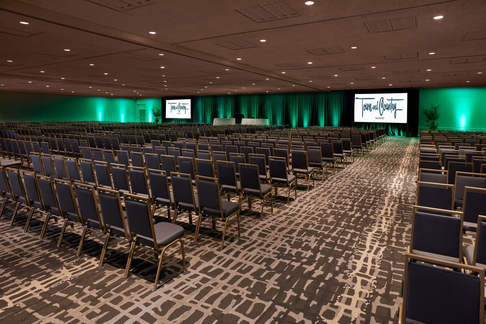 Large meeting hall with rows of chairs facing two screens under teal lighting
